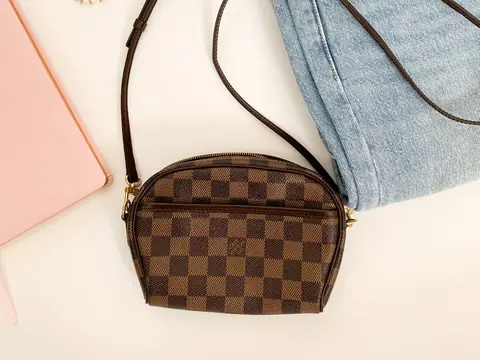 A Louis Vuitton Damier Ebene crossbody bag on a white table beside jeans and a pink notebook
