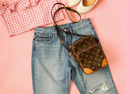A Louis Vuitton monogram canvas crossbody bag laid on jeans with a gingham top and cream shoes on a pink background